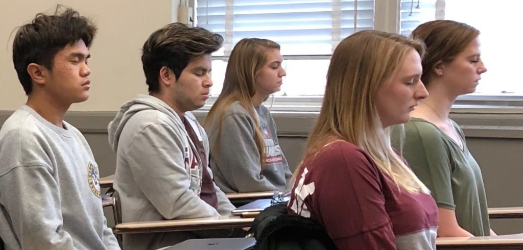 Students sit at desks with their eyes closed, meditating.