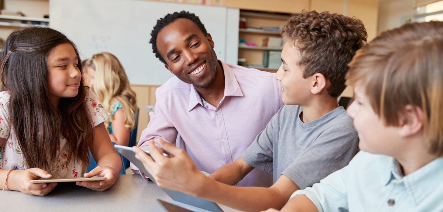 A teacher sits down at a table with a few students, engaging in conversation.