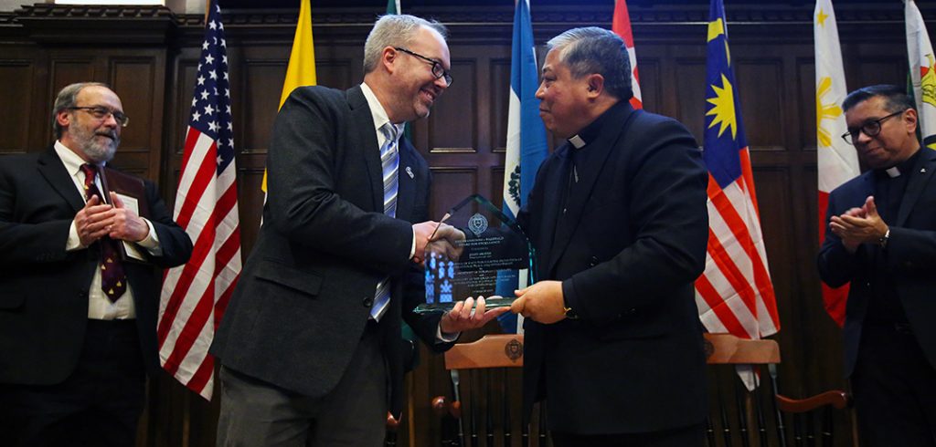 John Briggs shake hands with Archbishop Bernardito Auza on stage in the Keating First floor auditorium, with flags behind them.