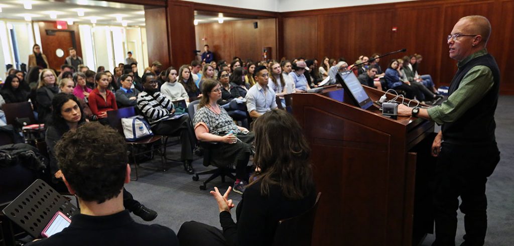 Eli Clare gives the 2019 Distinguished Lecture on Disability from a podium with interpreters nearby in a packed room