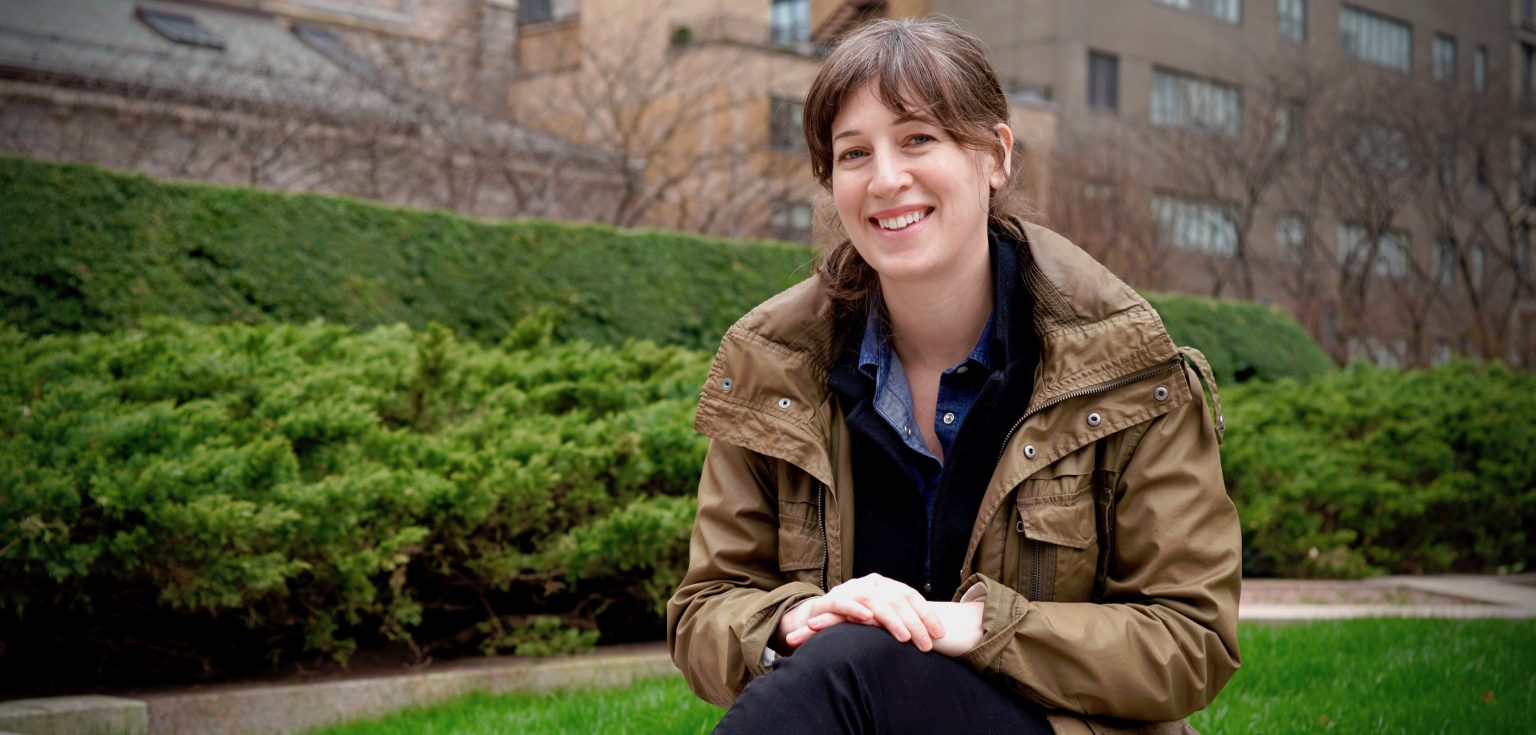 A brunette girl sits and smiles against a green and grassy backdrop.