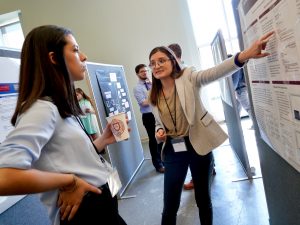 A girl wearing glasses and a blazer points toward her poster as another girl looks on.