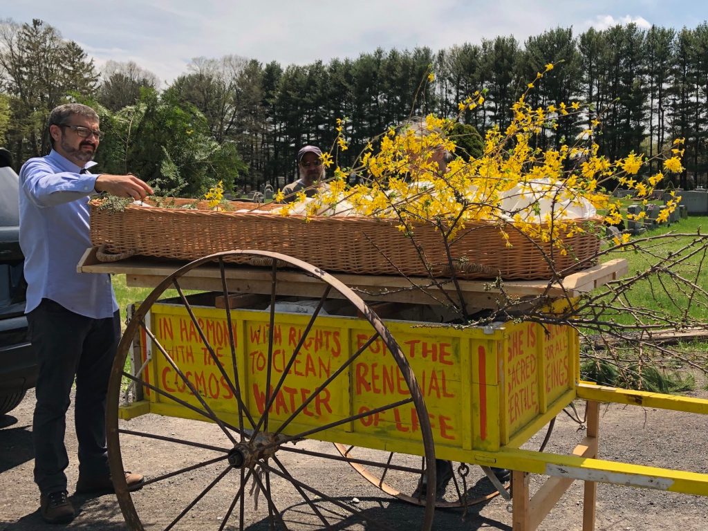 A wheelbarrow holding a wooden casket and a deceased person wrapped in a white shroud and yellow flowers