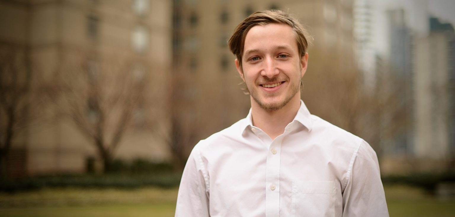 A blonde man wearing a white shirt and standing against a blurred city background