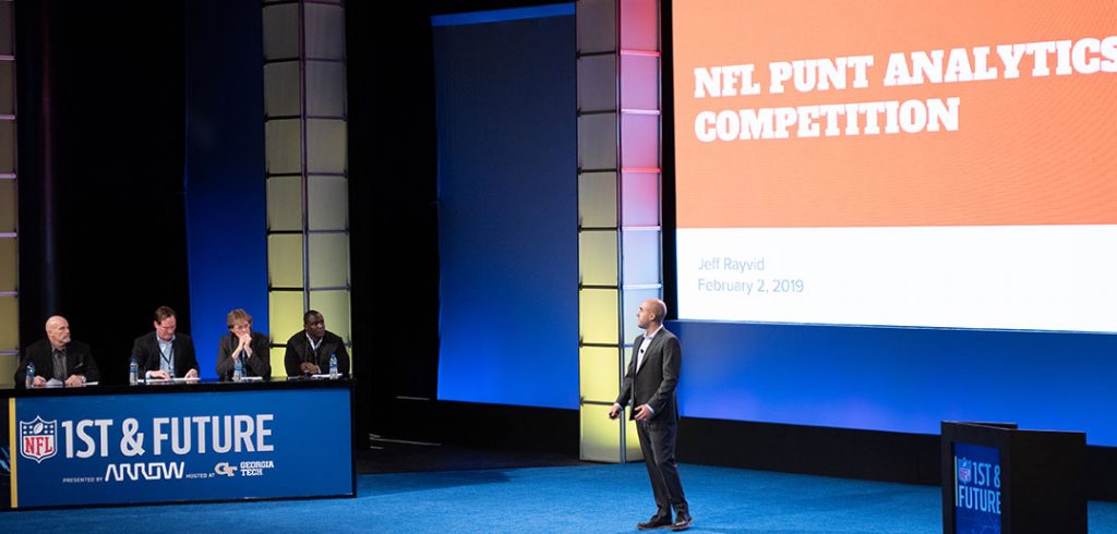 A student speaks from a large stage with a big screen behind him