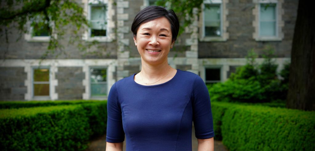 A smiling Asian woman wearing a blue dress and standing in front of a building