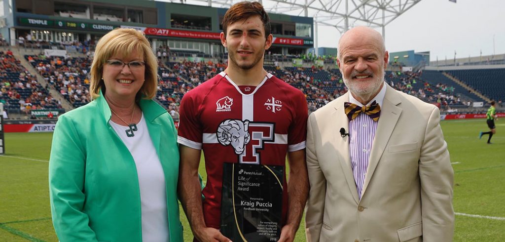 Eileen McDonnell, Kraig Puccia and Joe Jordan stand together on a field while Puccia holds a plaque