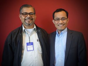 Two men wearing glasses and blazers smile at the camera against a red wall