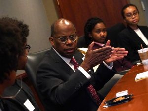 A man wearing a suit and glasses speaks and gestures with his hands.