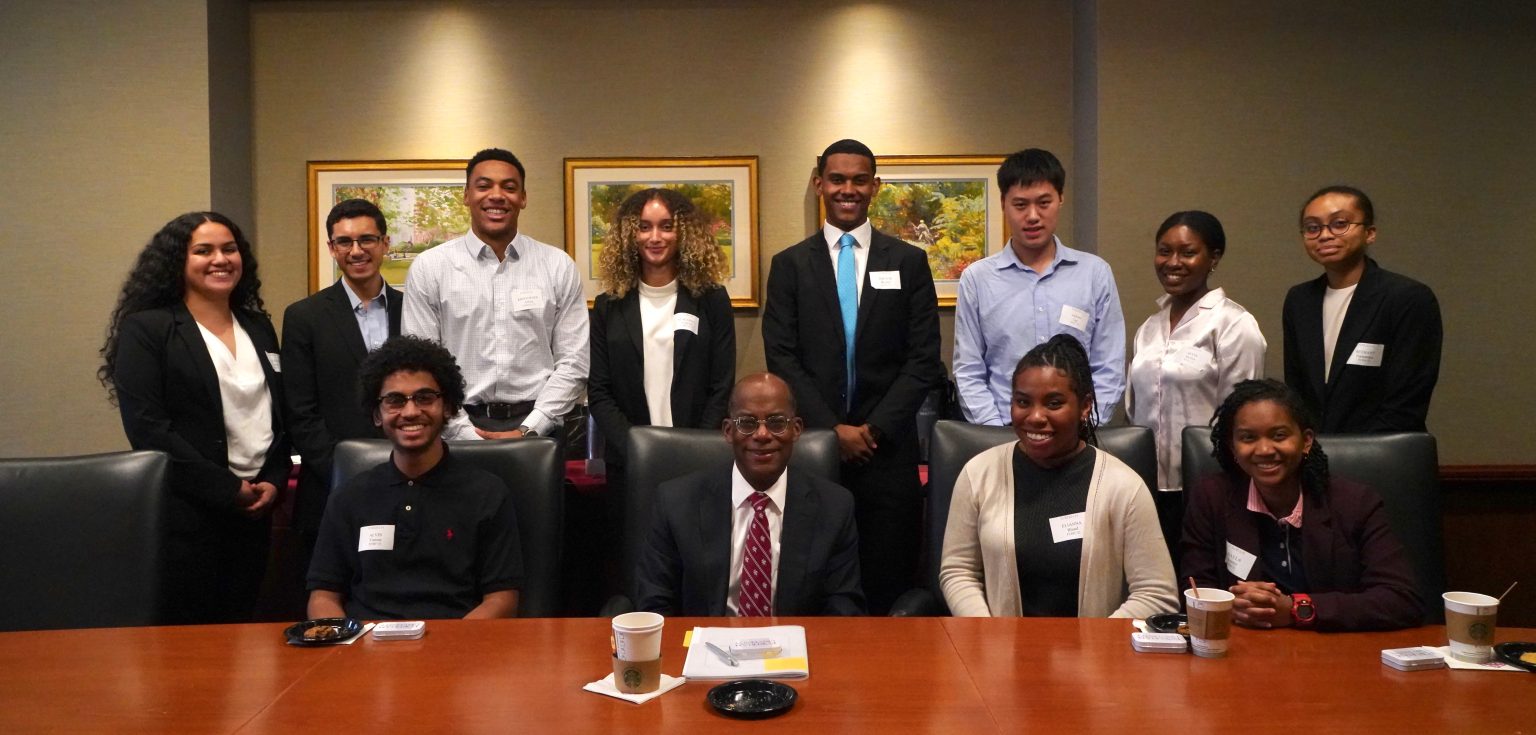 A group of students and a man wearing a suit pose for a picture in front of a big wooden table.