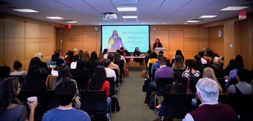 A large group of seated people looks at a PowerPoint presentation at the front of the room.