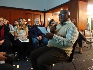 A maA man wearing glasses and a sweater vest sits and reads from a piece of paper.