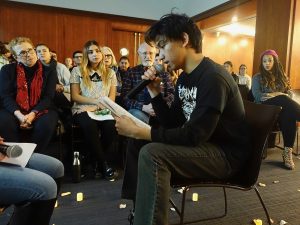 A young man wearing a black shirt sits and reads from a piece of paper.