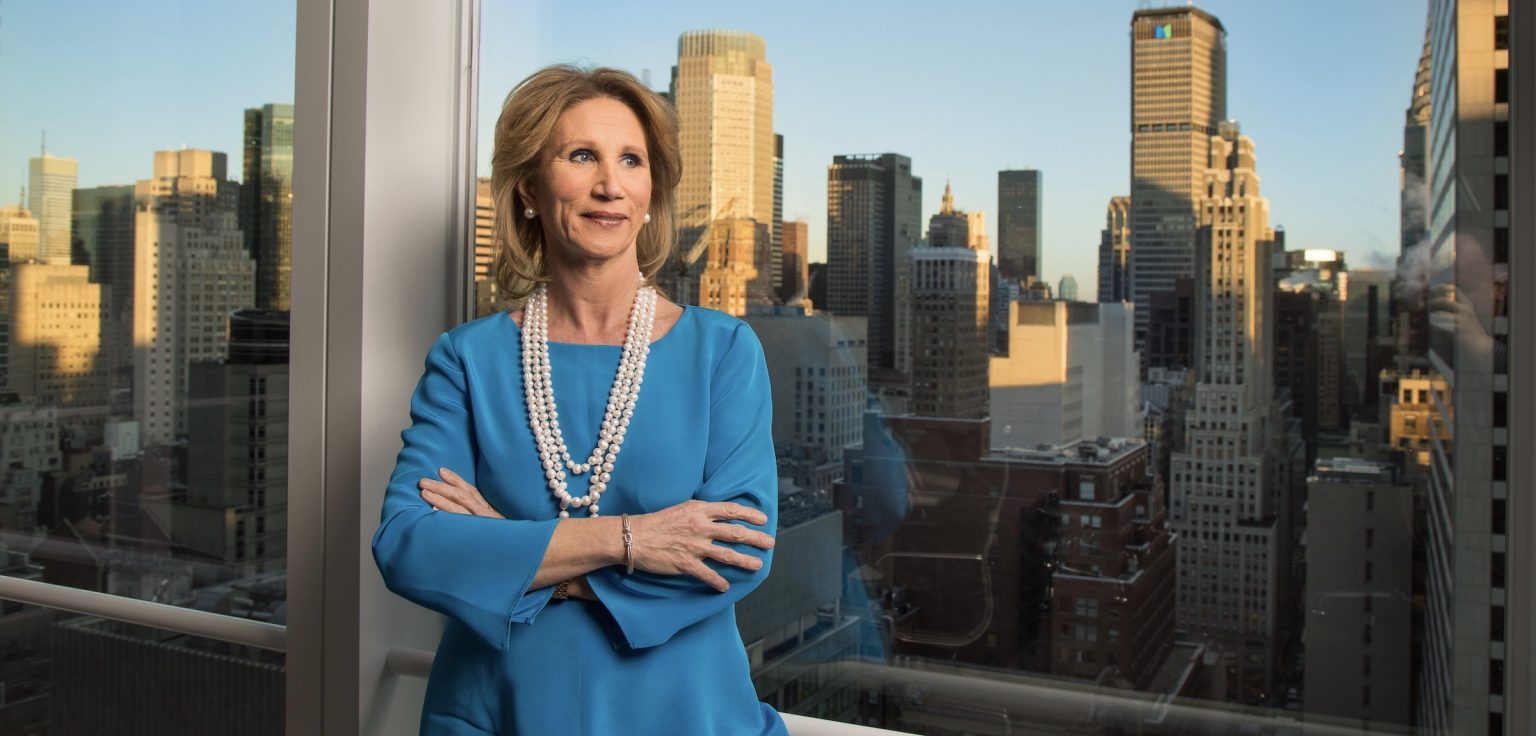 A woman wearing a blue shirt and long necklace stands in a building in front of the Manhattan skyline.