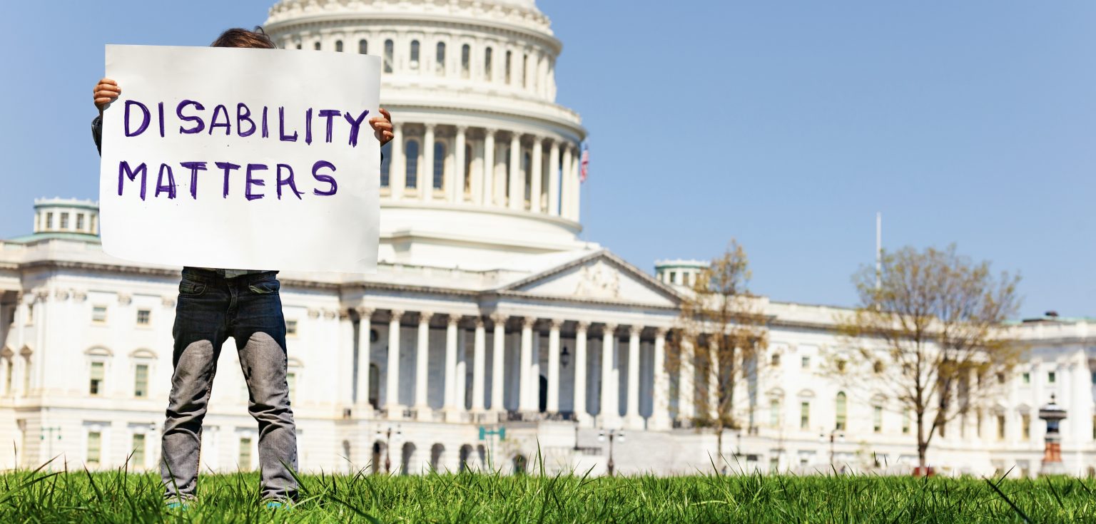 A boy stands in front of the Capitol Building and holds a handmade sign that says "DISABILITY MATTERS."