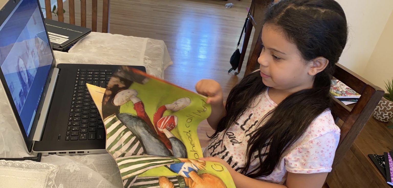 A little girl holds a book in front of an open laptop.