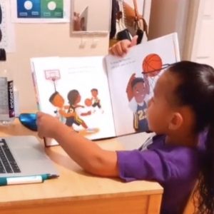 A little girl holds a book in front of an open laptop.