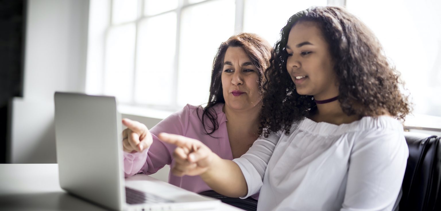 A middle-aged woman and a young woman sit in front of a laptop and point their fingers at it.