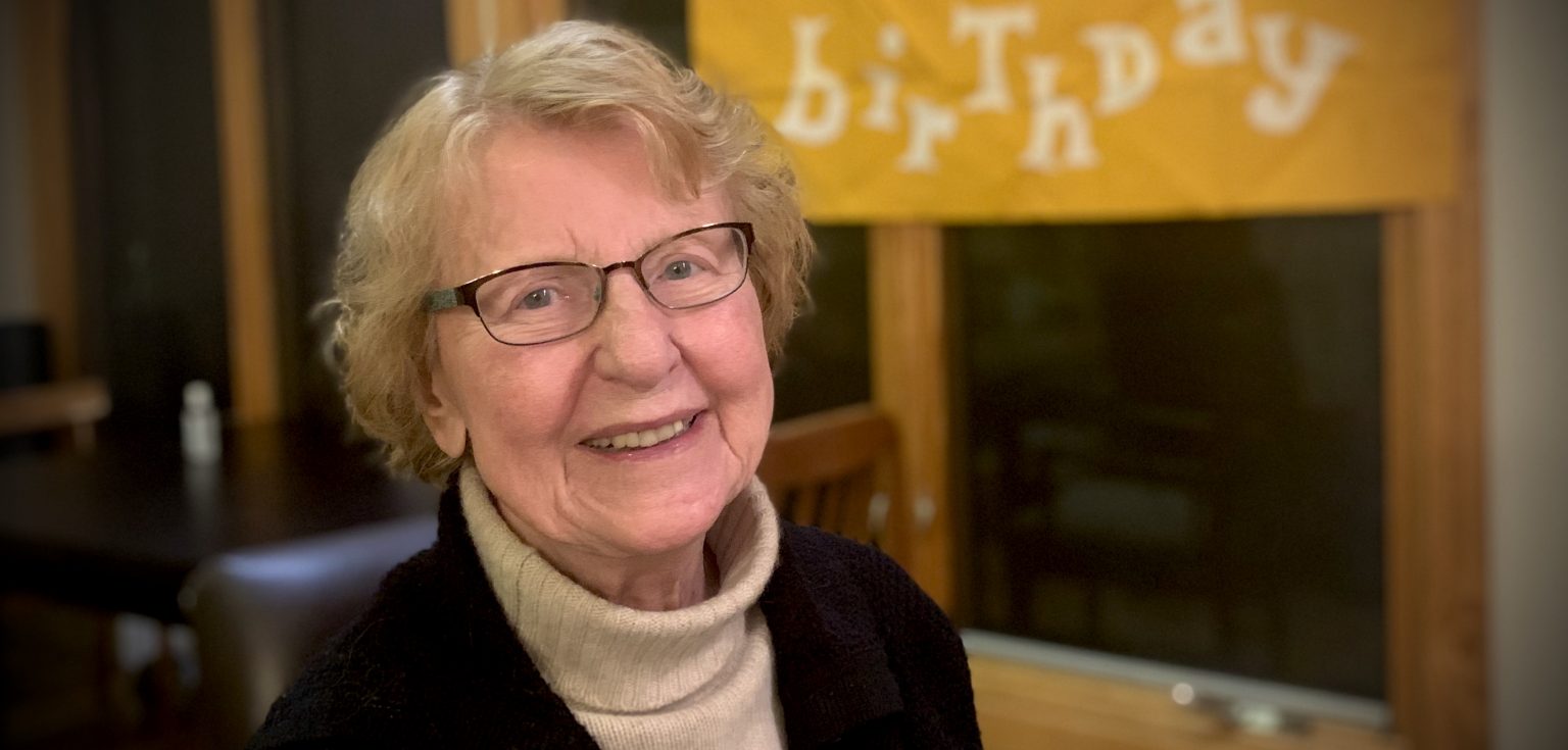 A white-haired woman wearing glasses and a beige turtleneck smiles at the camera.