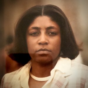 A woman with a somber expression stares at the camera, while wearing a necklace and a blazer.