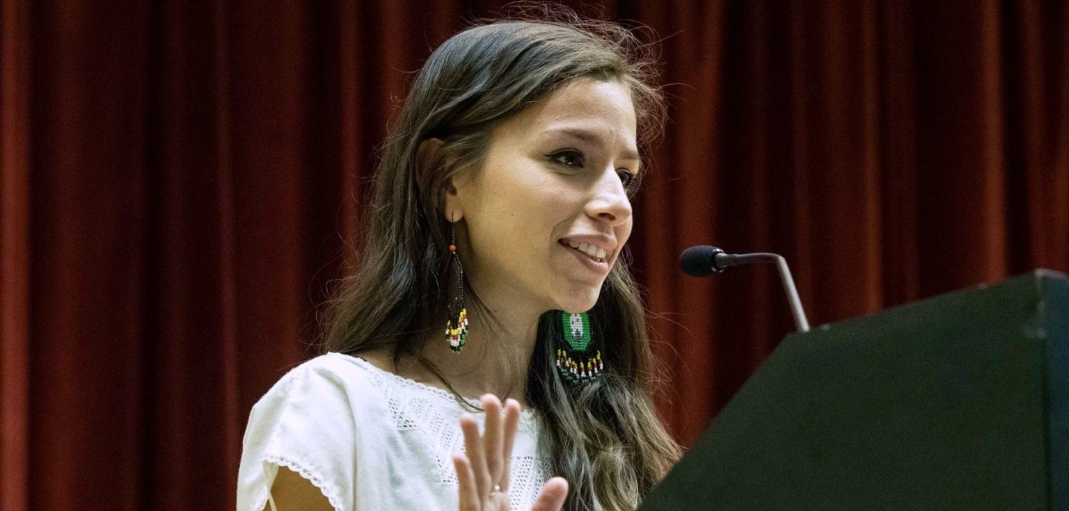 A woman with long hair speaks into a microphone at a podium.