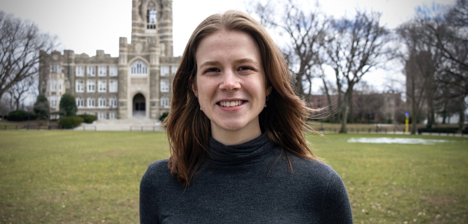 A brunette woman wearing a dark gray turtleneck smiles at the camera in front of a large green lawn.