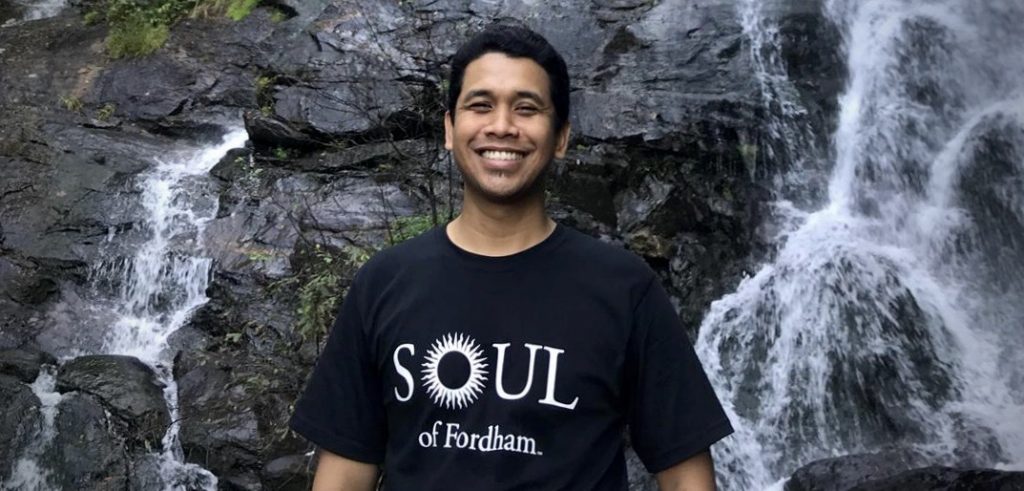 A man wearing a black t-shirt that says "Soul of Fordham" smiles in front of a waterfall.