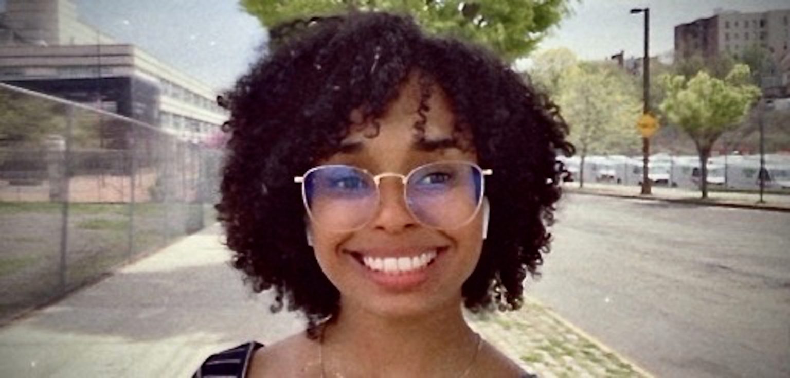 A woman with black curly hair and glasses smiles in front of a street.