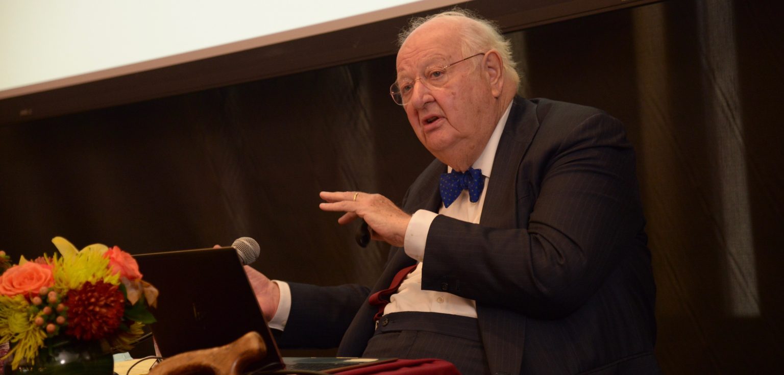 A seated elderly man wearing a suit and a blue bow speaks in front of a black curtain.