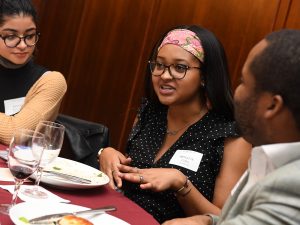 A woman with a headband and glasses speaks next to two people.