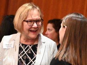 A woman with shoulder-length blonde hair, glasses, and a white blazer smiles at a student.