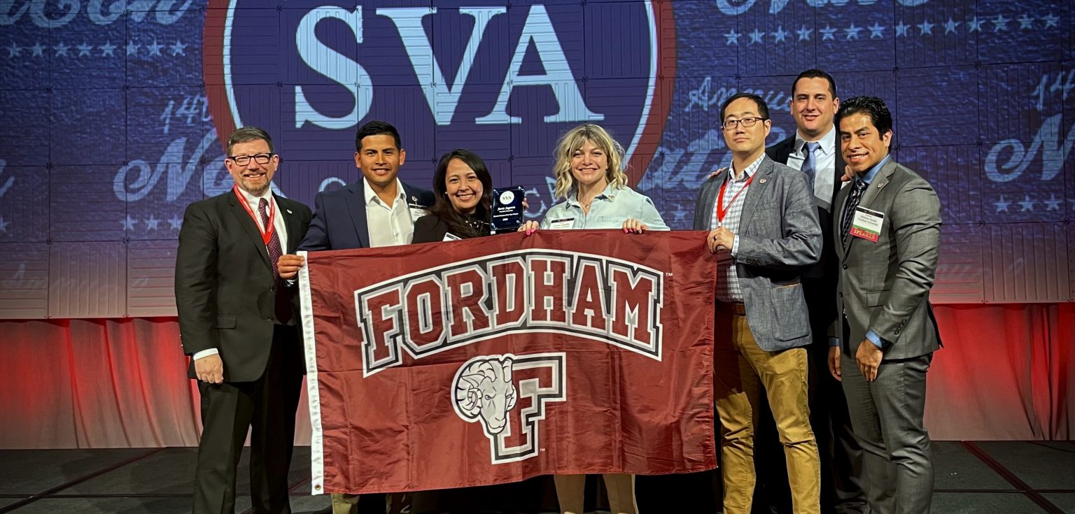 Seven people stand together and hold a maroon flag that says "Fordham."