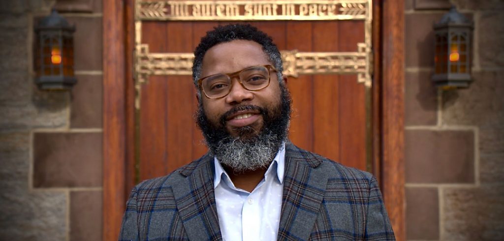 A man wearing glasses and a beard smiles in front of a church door.