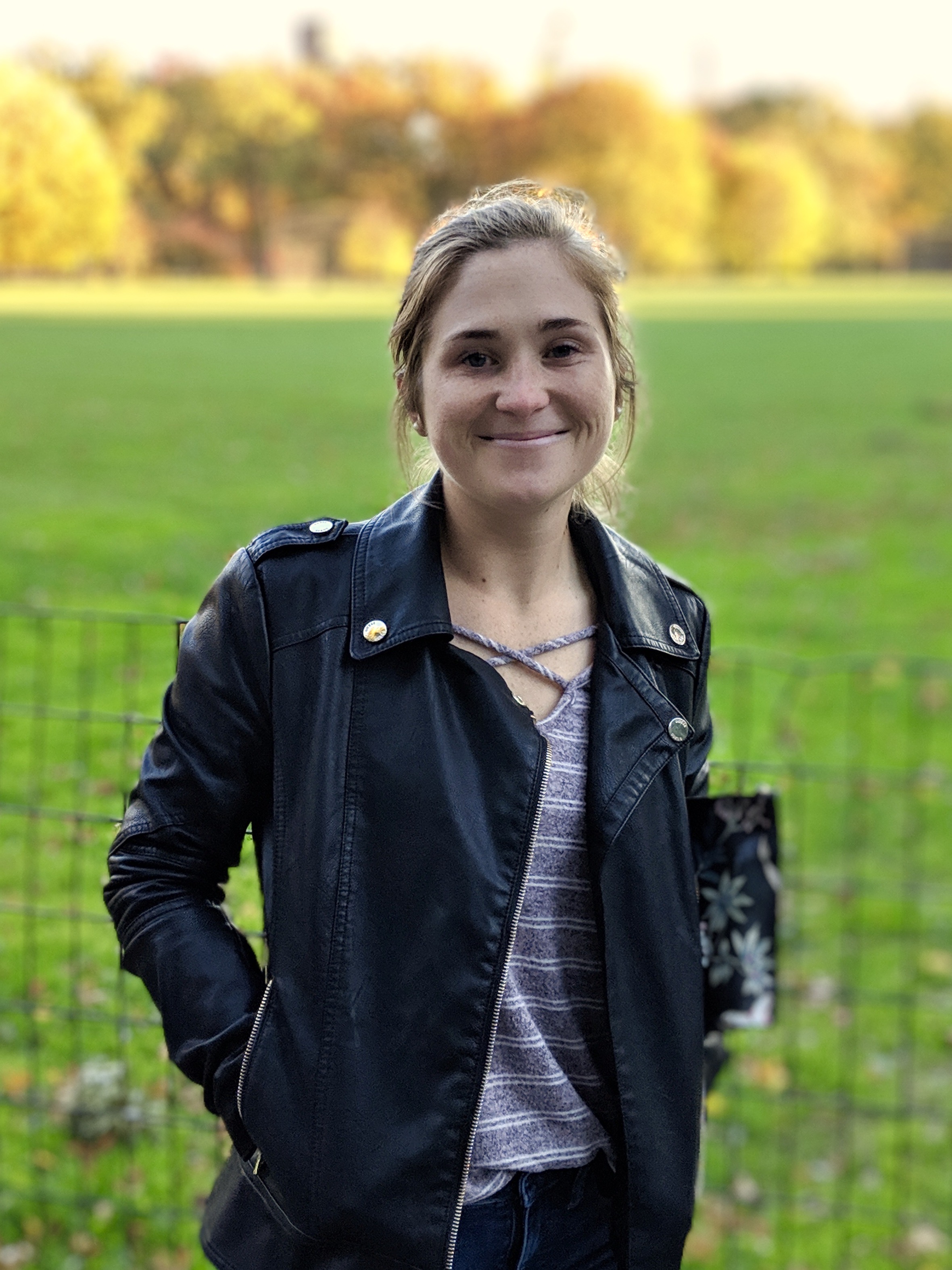 A woman wearing a black leather jacket smiles in front of a lot of grass.