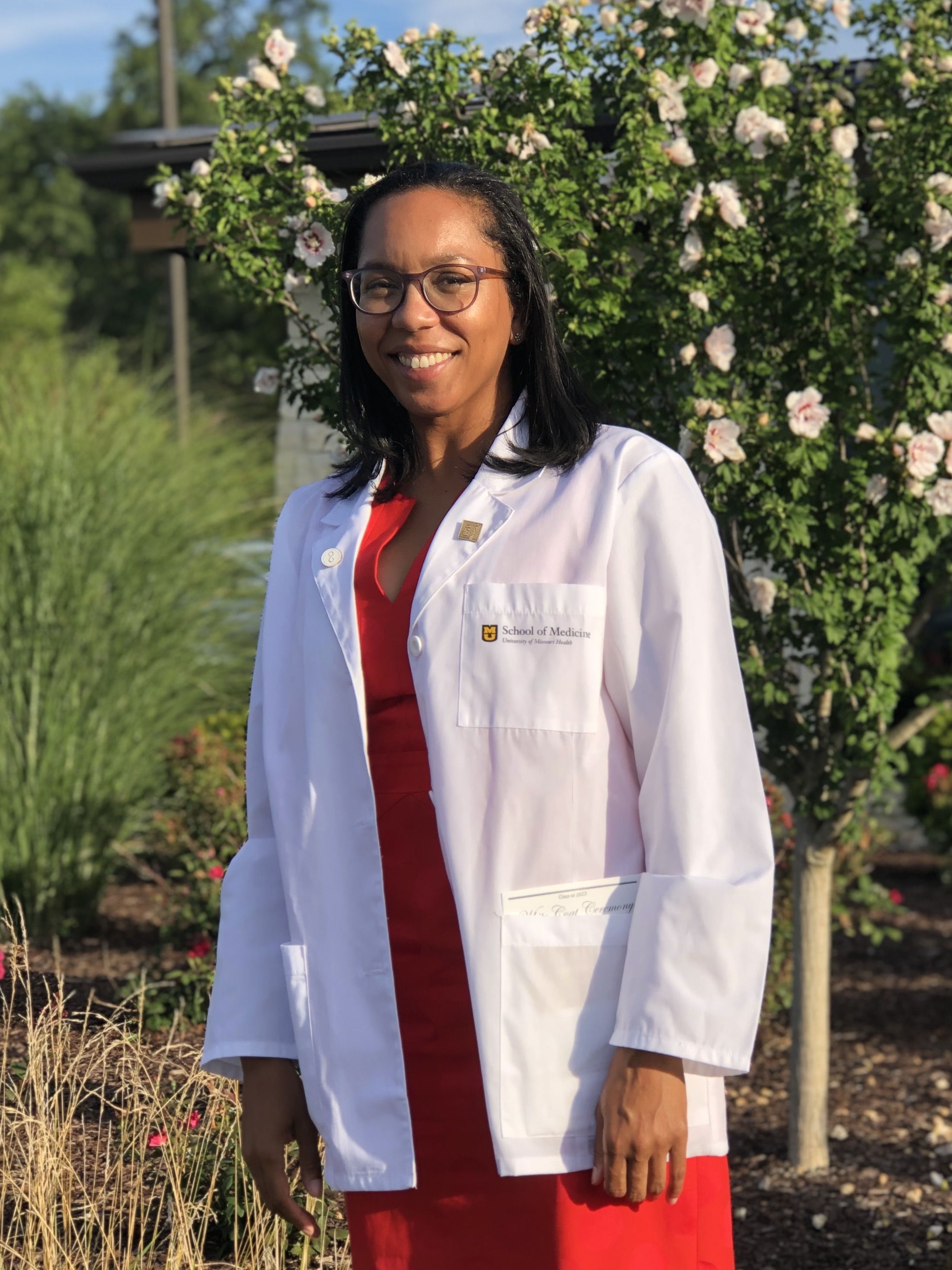 A woman wearing glasses, a red dress, and a white medical coat smiles in front of a grassy background.