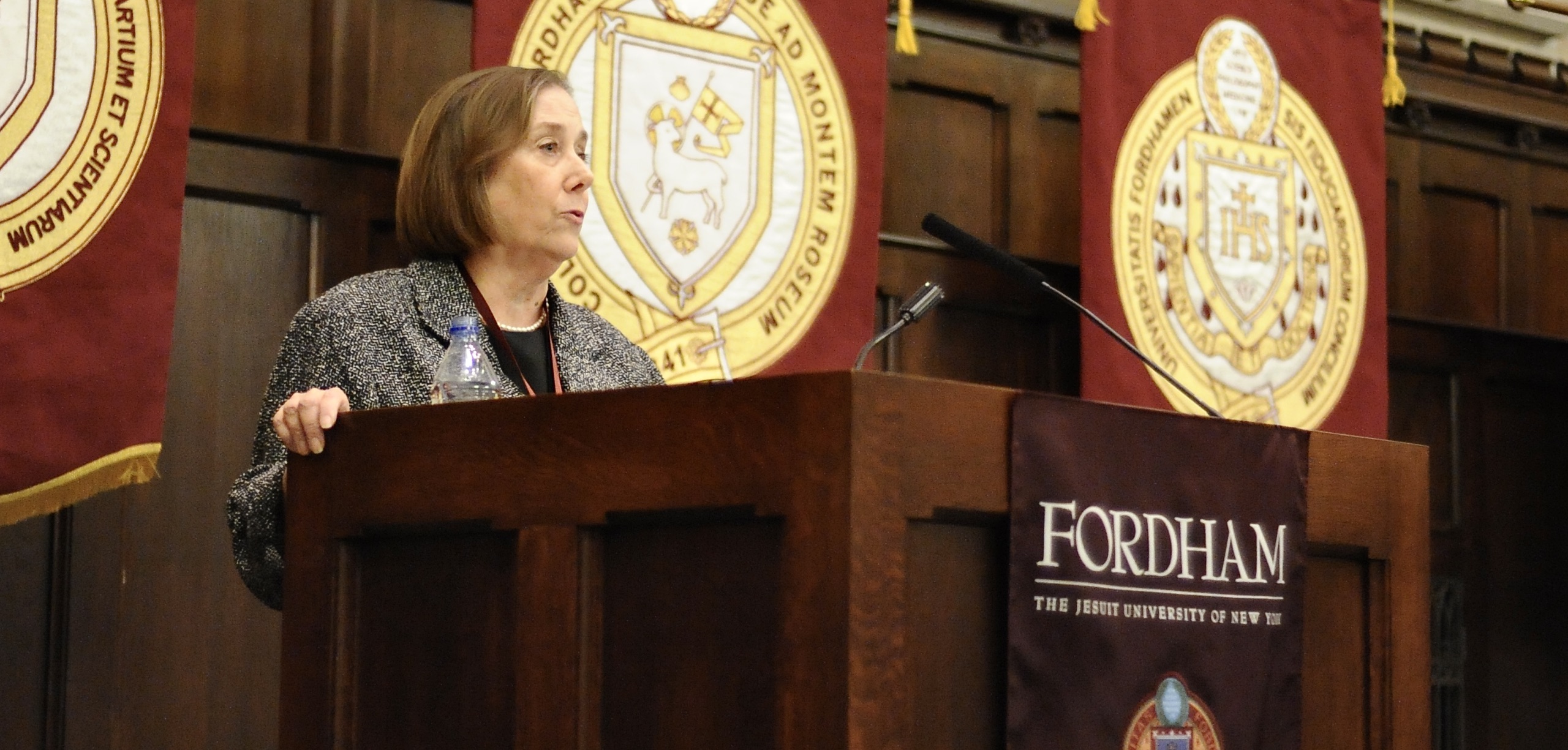 A woman stands at a wooden podium and speaks.