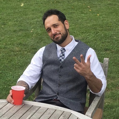 A man wearing a gray vest and white shirt holds a red solo cup and smiles.