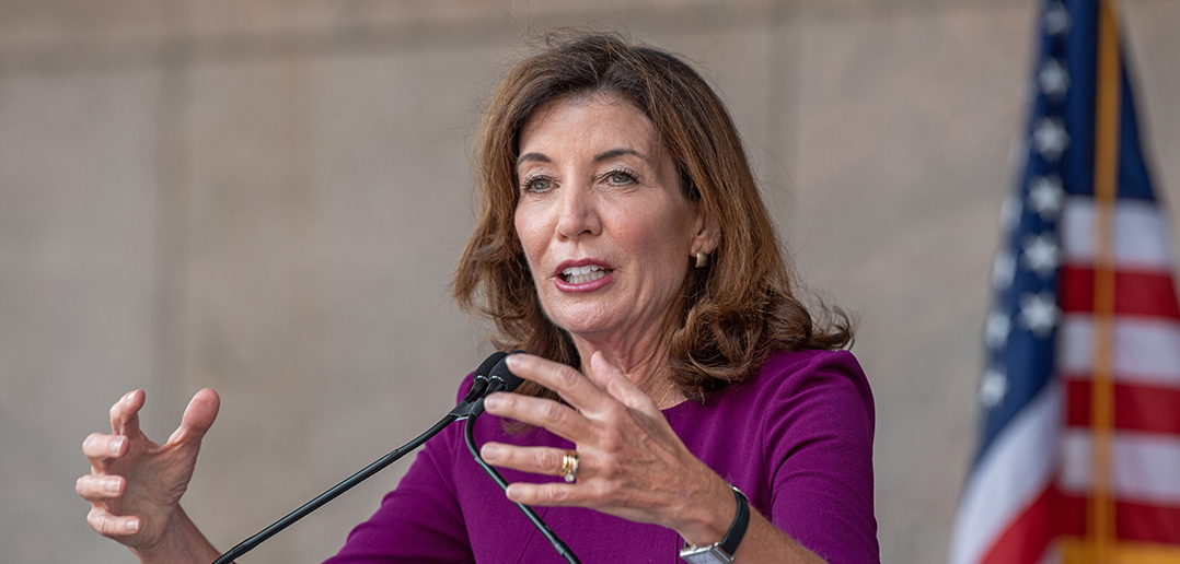 Kathy Hochul talking with her hands at a podium with the American flag to her left