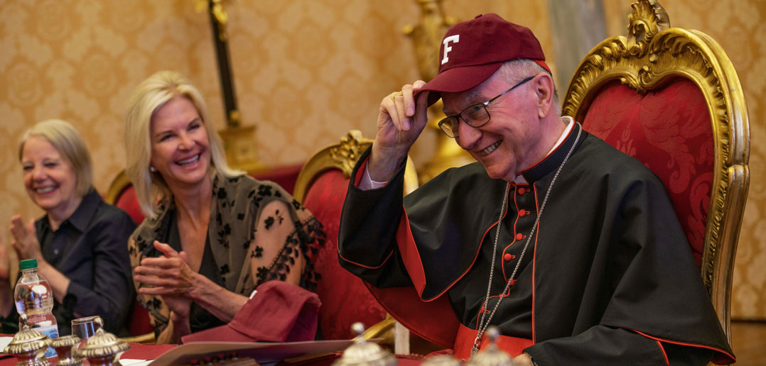 A man wearing a maroon baseball cap and a black priestly outfit smiles.