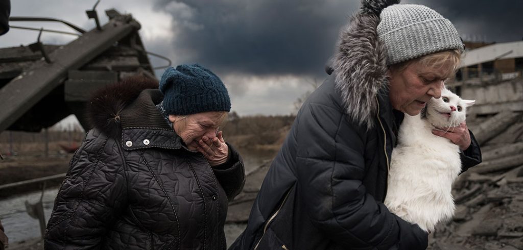 Two women, one holding a white cat in her arms, the other covering her mouth with her hand, flee a battle scene in Irpin, Ukraine