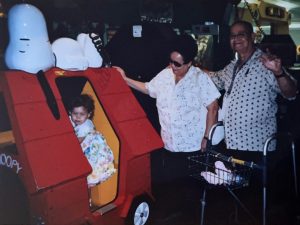 An elderly couple stands by a little girl who is standing in a red playhouse.