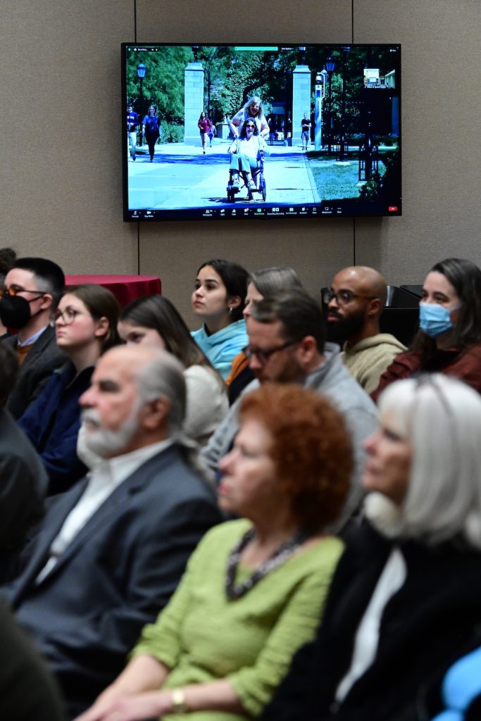 People face forward and listen to an unpictured speaker with a TV in the background that features a person and a second person in a wheelchair.