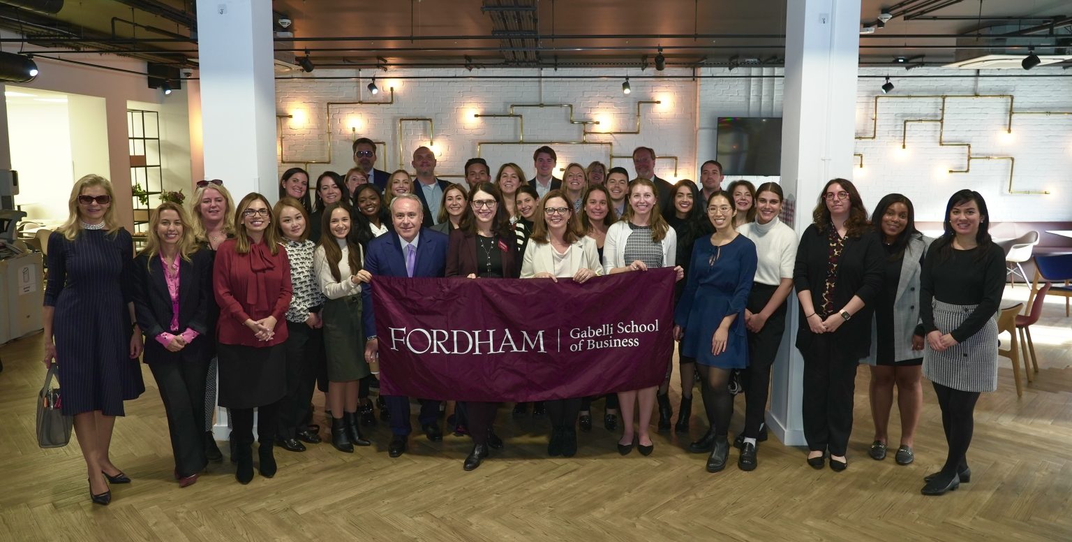 A large group of people stand and smile while holding a maroon flag that says "FORDHAM Gabelli School Business."