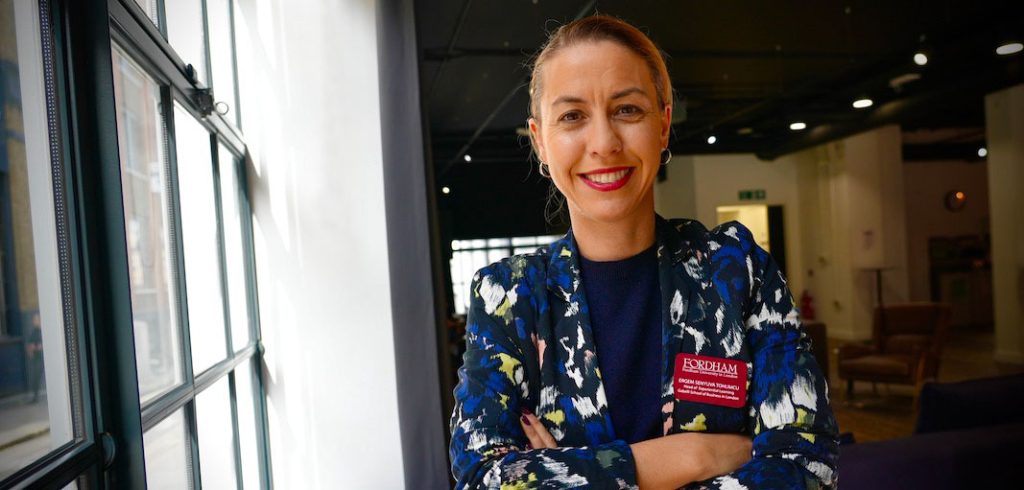 A woman wearing a floral suit jacket crosses her arms and smiles beside a window.