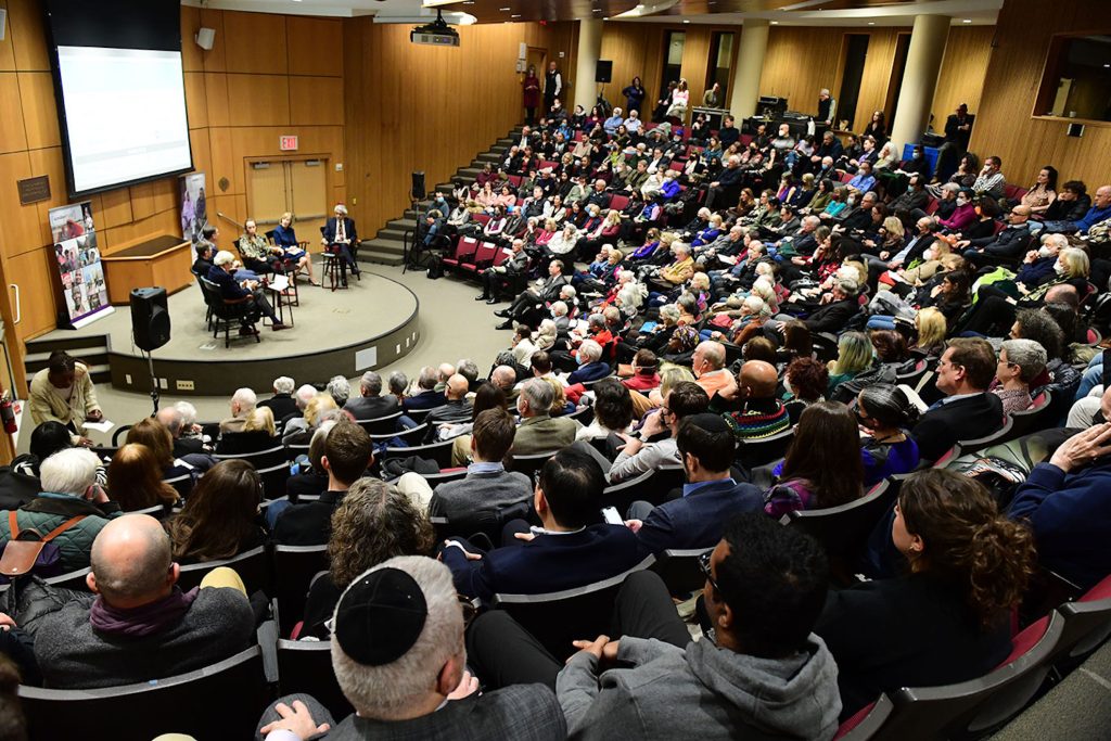 A crowded auditorium of people listens to a group of speakers sitting on an elevated platform.