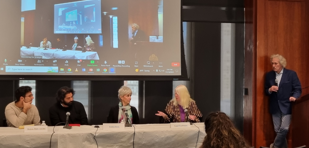 Two men and two women on a panel. There is a projector screen behind them. There is a man standing to their left.