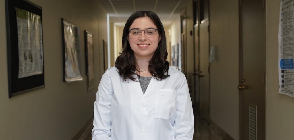 A woman wearing a white lab coat smiles in a hallway.