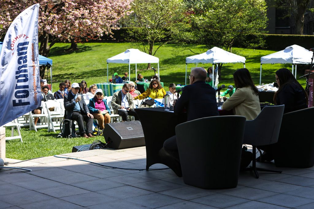 Several people seated, with an audience arrayed out on a lawn in front of them