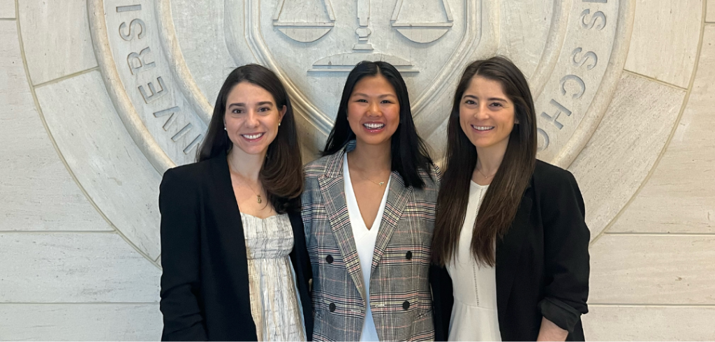 Bianca Bernardi ’23, Millicent Kastenbaum ’23, and Sofia La Bella ’23, standing together dressed in professional attire. They are behind a wall with the Fordham seal on it.