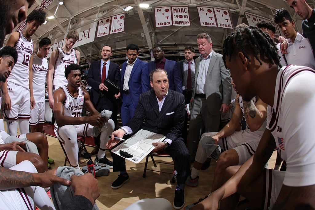 Keith Urgo sitts in the middle of the mens basketball team on the court, holding a clip board.